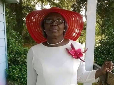 Azalee standing outside wearing a white dress, red corsage and matching red wide brim hat.