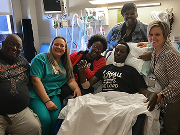 DeAndre wearing a black tee-shirt and sitting in bed, surrounded by his family and members of his care team.
