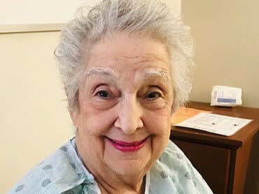 Elaine smiles sitting in a chair in her hospital room.