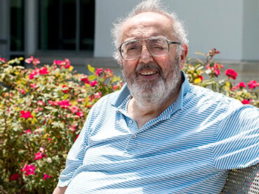 Bill sits on a bench in a rose garden on a sunny day.