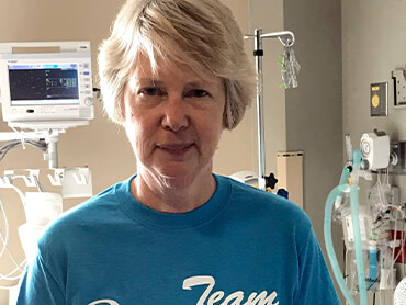 Sharon standing in hospital room wearing a blue tee-shirt with a vital signs monitor in background.