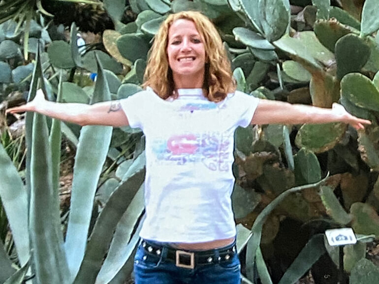 Tara smiling, arms outstretched, standing in an ornamental garden.