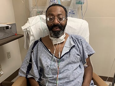 Veron sitting in a chair in his hospital room with IV equipment in the background.