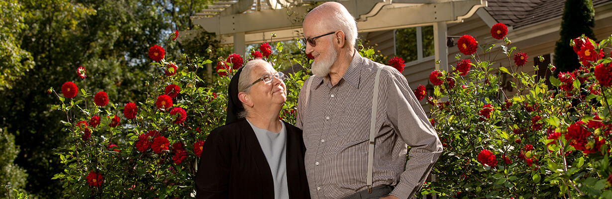 Smiling elderly couple standing among red flowers in a garden, looking at each other.